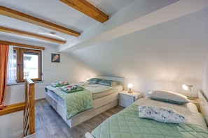 A bedroom with two beds, wooden beams, a window with curtains, a bedside table with a lamp, and a wall-mounted picture.