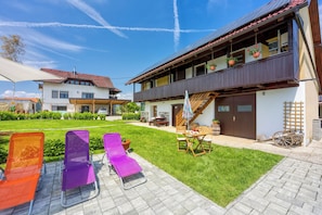 A two-story building with a balcony, a wooden table with chairs, and a garden area with lounge chairs.