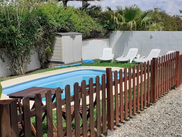 A swimming pool with a wooden deck and a fence, surrounded by greenery and a white wall.