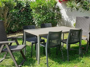 Mesa rectangular al aire libre con cuatro sillas, rodeada de abundante follaje y un muro de ladrillo.