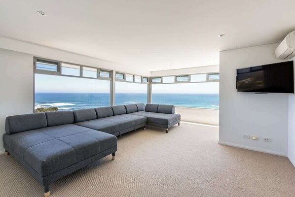 Una sala moderna con un amplio sofá seccional gris, un televisor de pantalla plana montado en la pared y un ventanal con vistas al océano.