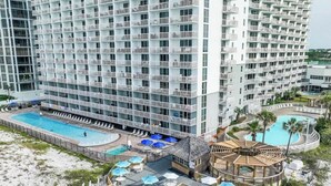 Un hotel moderno de gran altura con varios balcones, un área de piscina y un lounge frente a la playa con sombrillas.