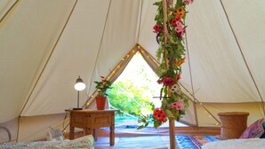 Una carpa con dos camas, una mesita con una lámpara, una planta en maceta y un arreglo floral colgante.
