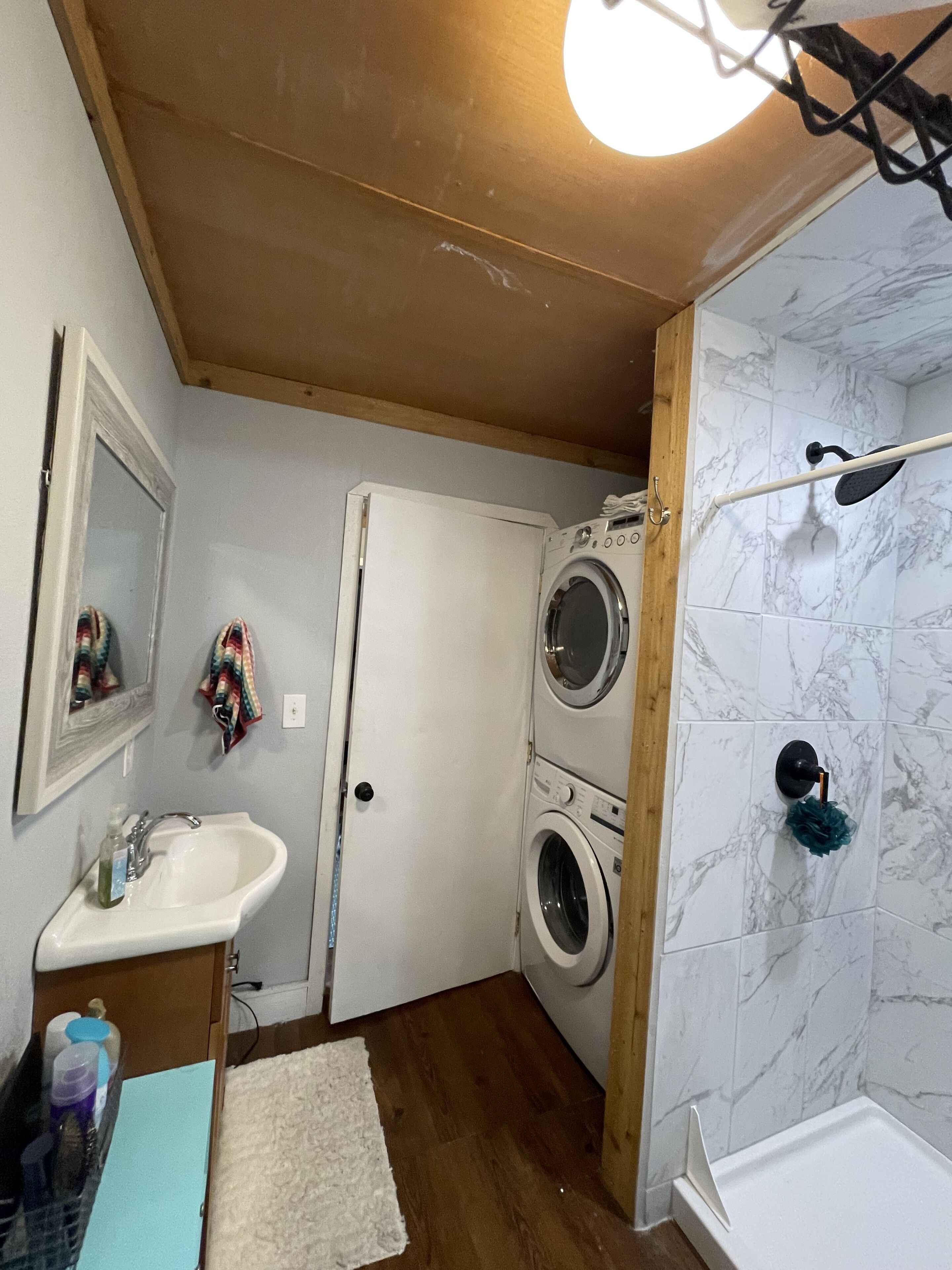 A compact bathroom with a sink, two stacked washing machines, a shower area, and a mirror.