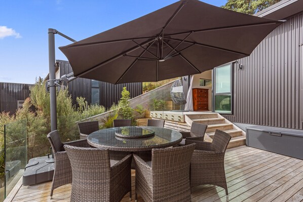A modern outdoor patio with a glass table surrounded by wicker chairs, a large umbrella, and a wooden deck.