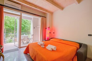 Habitación de hotel con colcha naranja, puerta corrediza de vidrio a un balcón y una mesa con sillas afuera.