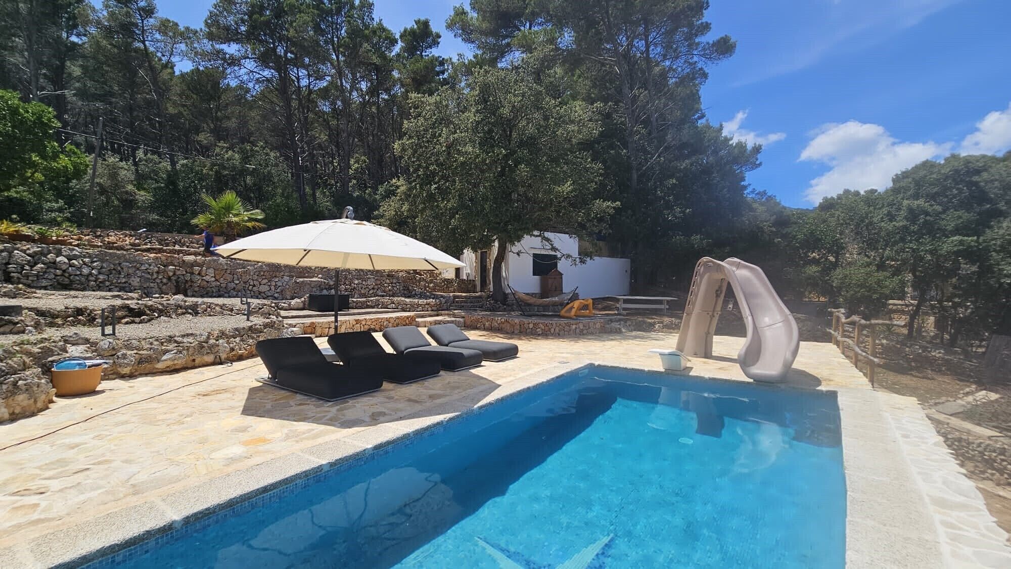Une piscine dotée de chaises longues et d’un toboggan, entourée de murs en pierre et d’arbres.