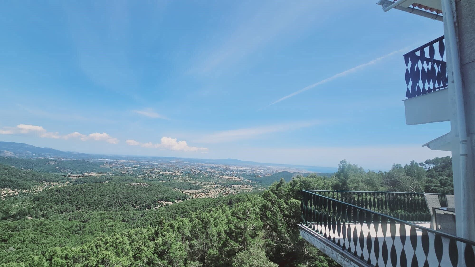 Un balcon offrant une vue sur une vallée densément boisée et sur des montagnes lointaines, le tout sous un ciel dégagé.