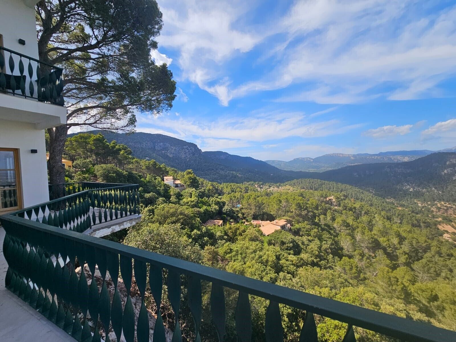 A balcony with a green railing overlooking a mountainous landscape with trees and buildings.