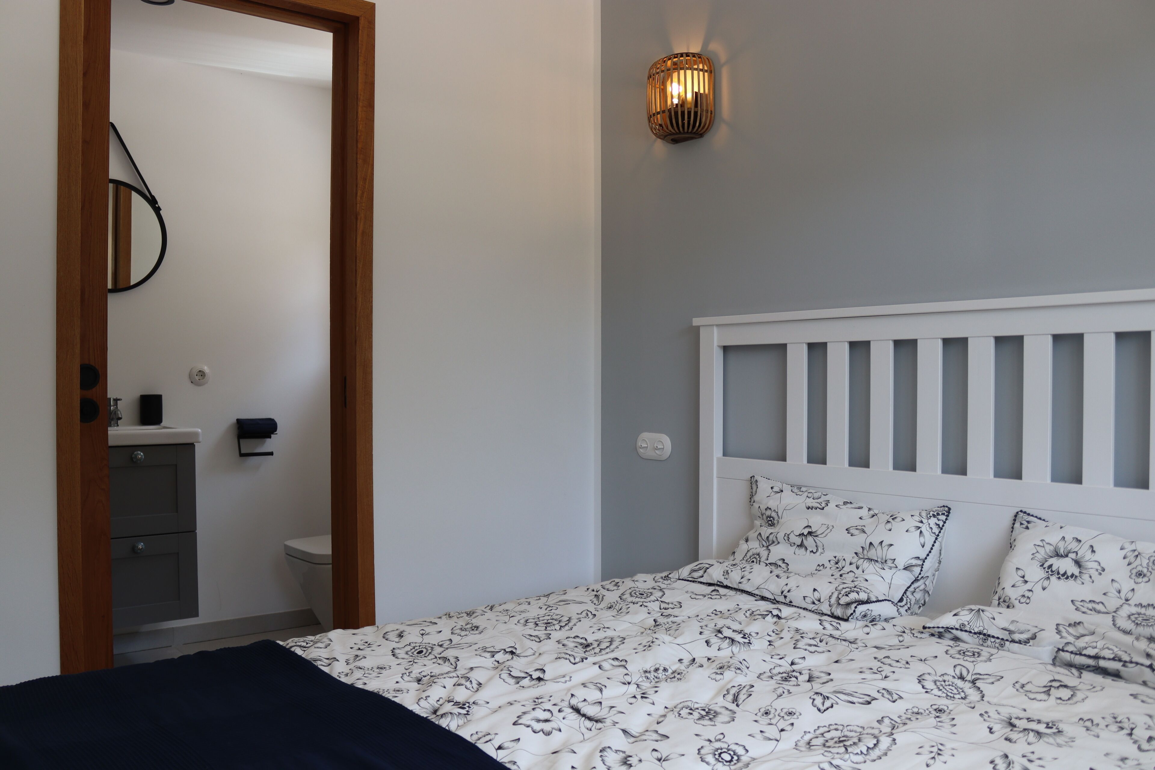 A bedroom with a white headboard, floral bedding, a bedside lamp, and a view into the bathroom.
