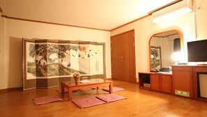 Ein traditioneller japanischer Raum mit Tatami-Böden, einem niedrigen Holztisch, einem Faltbildschirm mit Kunstwerk, einem Fernseher und einem Schrank.
