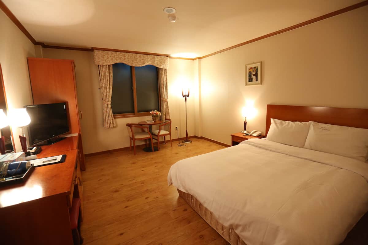 A hotel room with a wooden desk, a television, a bed with white linens, a desk lamp, and a window with curtains.