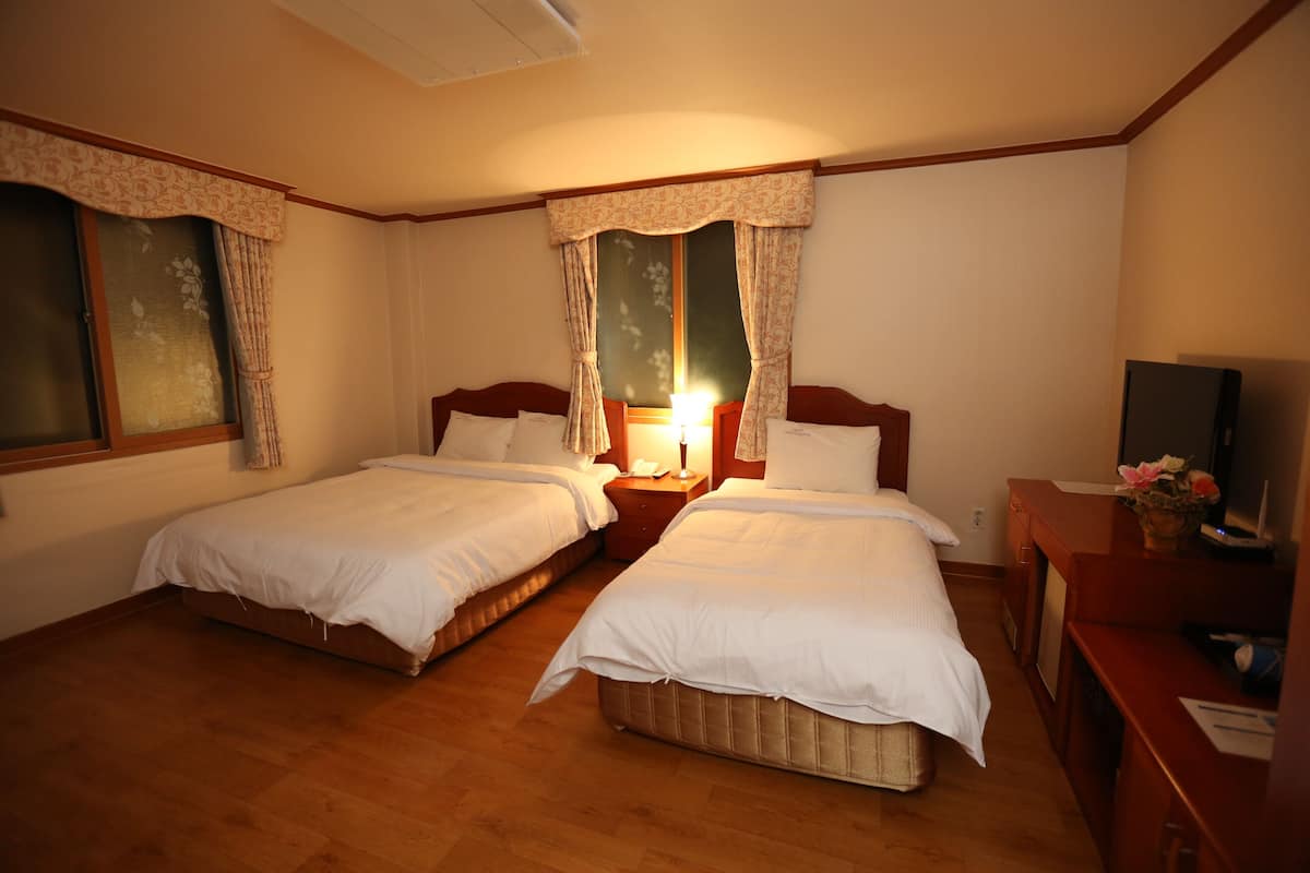 A hotel room with two single beds, wooden flooring, a desk with a chair, a television, and a window with curtains.