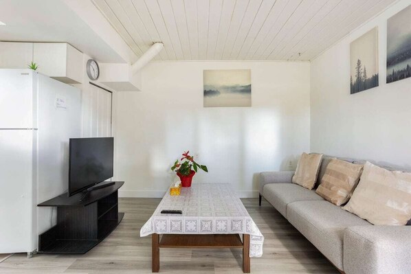 Ein modernes Wohnzimmer mit einem Flachbildfernseher auf einem schwarzen Standfuß, einem weißen Tisch mit Spitze-Tischdecke, einem grauen Sofa mit beige farbenen Kissen und einem Gemälde an der Wand.