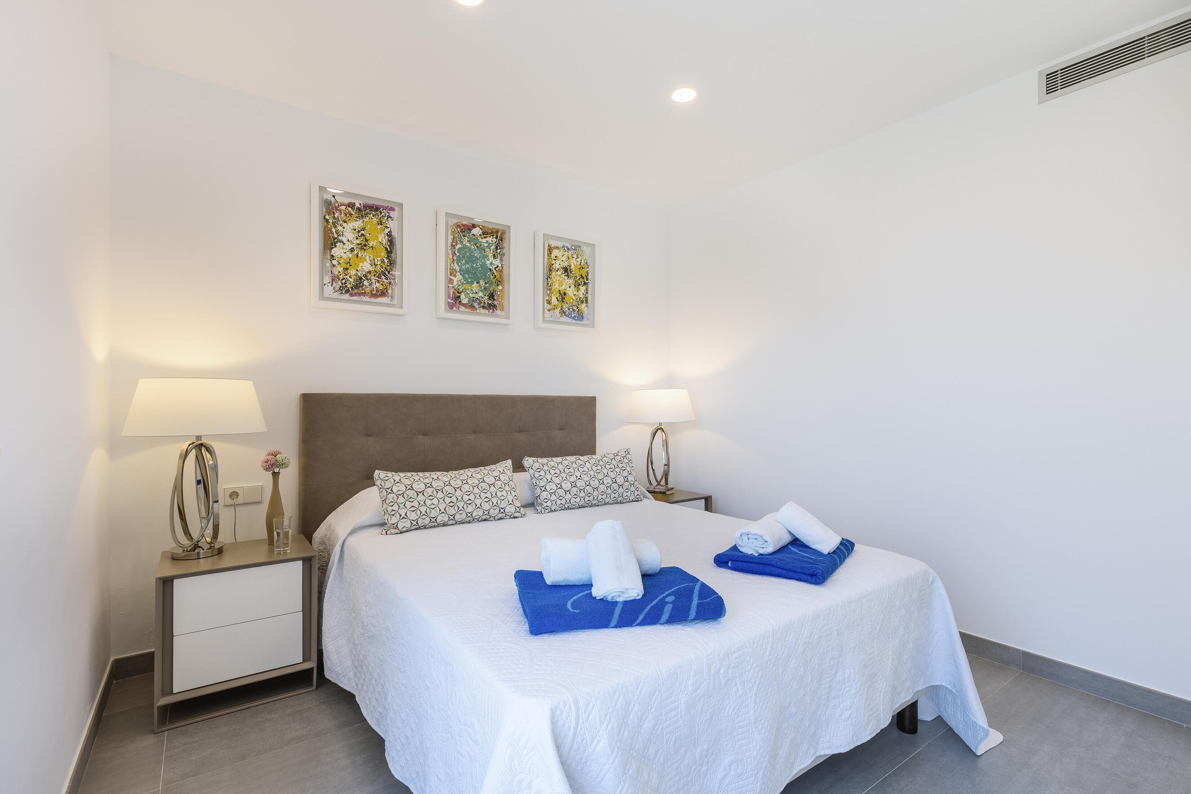 A neatly made bed with white and blue linens, a bedside table with a lamp, and three framed artworks on the wall.