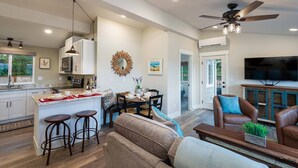 A modern kitchen with white cabinets, a dining area with a round table, and a living room with a brown leather couch and a TV.