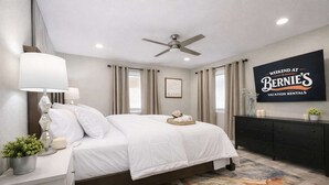 Una habitación de hotel con una cama, mesitas de noche, un ventilador de techo, un ventanal con cortinas, un televisor montado en la pared y una alfombra decorativa.