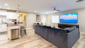 Un espacio habitable moderno con cocina, un sofá seccional gris, televisor de pantalla plana y ventilador de techo.