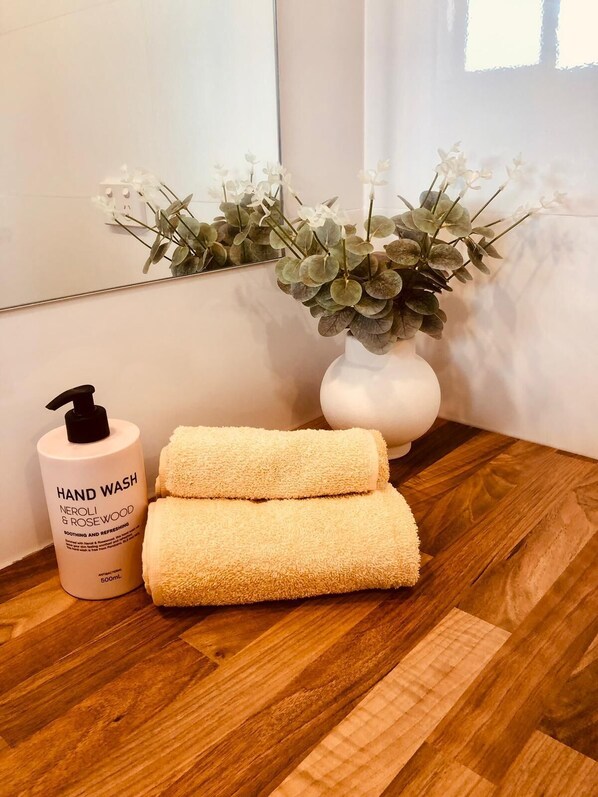A hand wash bottle, two rolled towels, and a vase with flowers on a wooden floor. - Shoalhaven Executive Apartments (Nowra)