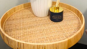 Una mesa redonda de madera con superficie trenzada, una maceta de cerámica blanca con una planta y un recipiente negro con tapa dorada y difusor de cañas.