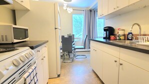 Ett kompakt kök med mikrovågsugn, spis och diskho. Det finns en matplats med ett bord och stolar som syns genom en öppen dörr.