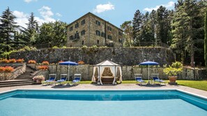 Un área de piscina con sillones de descanso y sombrillas, un edificio de piedra y un muro de piedra con escaleras que suben a él.