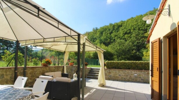 An outdoor patio with a covered seating area, stone walls, and a view of a forested hill. - MULINO DELLA RASSINA - MULINO DELLA RASSINA (Arezzo)