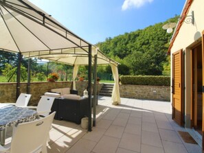 An outdoor patio with a covered seating area, stone walls, and a view of a forested hill.