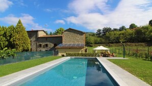 A rectangular swimming pool with a stone border, surrounded by a well-maintained lawn and a stone building with solar panels in the background. - MULINO DELLA RASSINA - MULINO DELLA RASSINA (Arezzo)
