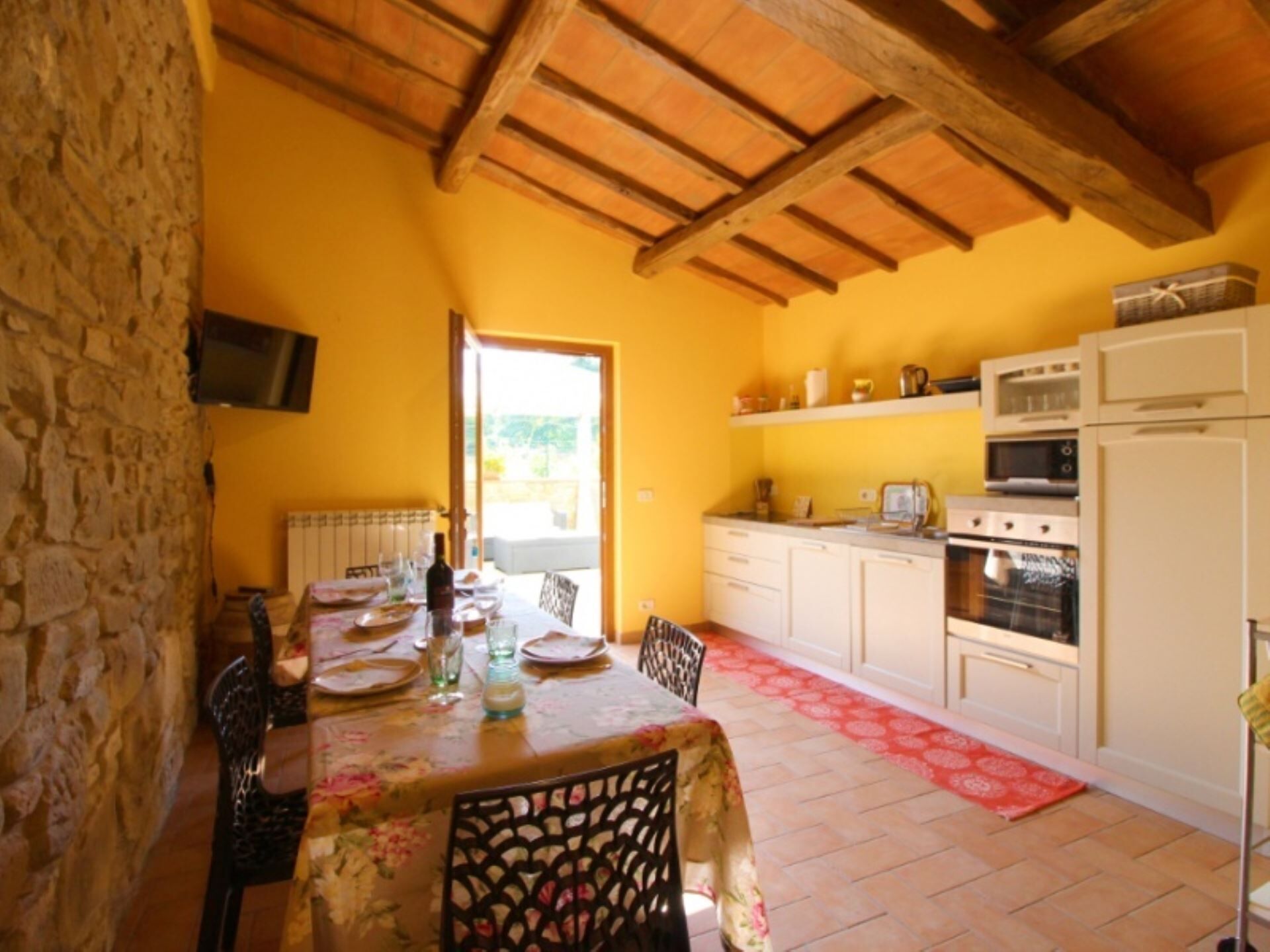Zona pranzo con tavolo apparecchiato per quattro, parete in pietra e cucina con mobili ed elettrodomestici bianchi.