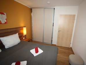 A hotel room with a bed, a nightstand with a lamp, a wardrobe, and a door. - HLI 3.10 up to 6 people 61sqm - Residenz Hohe Lith Cuxhaven (3.1.1) (Cuxhaven)