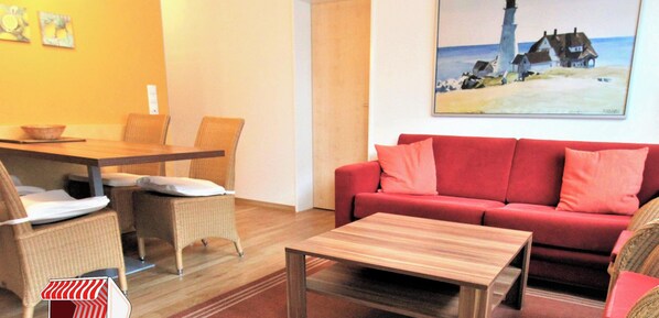 Uma sala de estar com um sofá vermelho, uma mesa de centro de madeira, cadeiras de vime e um quadro com uma cena de praia.