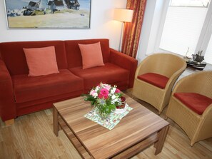 Um sofá vermelho, duas cadeiras de vime, uma mesa de centro de madeira com um arranjo floral, um abajur e um quadro na parede.