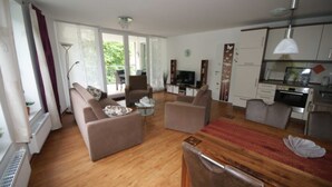 A spacious living room with a wooden floor, two sofas, a dining table, and a kitchen area with a TV and cabinets. - HLI 3.13 4 persons 77sqm - Residence Hohe Lith Cuxhaven (3.1.1) (Cuxhaven)