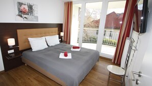 Quarto de hotel com uma cama grande, mesa de cabeceira com abajur, um quadro na parede e uma varanda com cortinas.