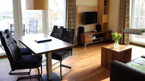Un salon moderne avec une table à manger en bois, des chaises noires, un téléviseur à écran plat, un meuble en bois et une petite plante posée sur une table d’appoint.