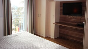 Une chambre à coucher avec un lit, une armoire, un téléviseur posé sur un meuble en bois et une fenêtre avec des rideaux.