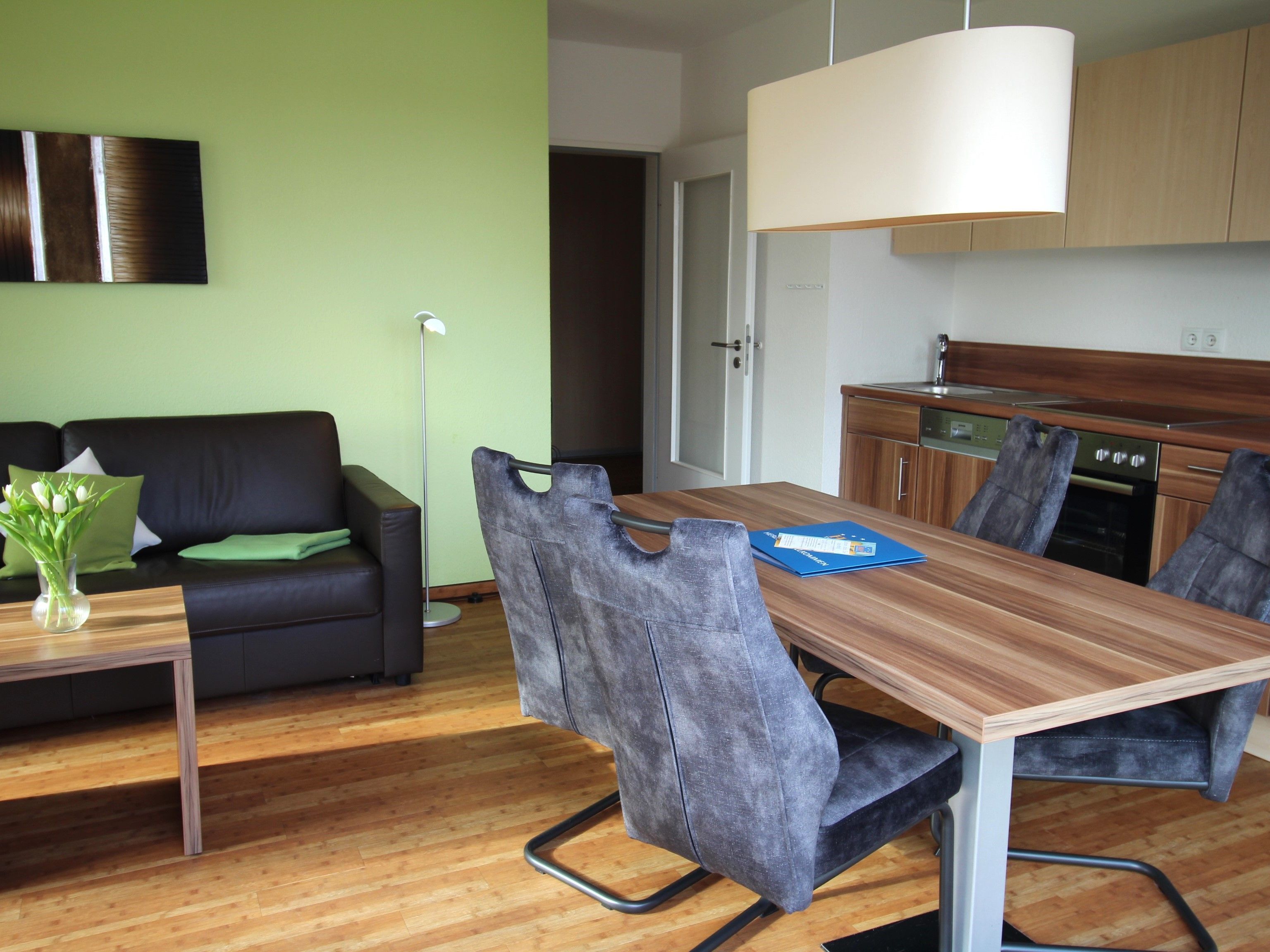 Un salon moderne avec un canapé noir, des coussins verts, une table basse en bois et un vase de fleurs. À côté se trouve un coin repas avec une table en bois, des chaises grises et un classeur bleu. On aperçoit à l’arrière-plan une cuisine avec des armoires en bois et un évier en acier inoxydable.
