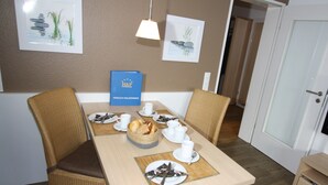 Ein Essbereich mit einem Tisch für vier Personen, gedeckt mit Tellern, Besteck und einem Brotkorb. Es sind Korbstühle und eine blaue Willkommensstasche auf dem Tisch.