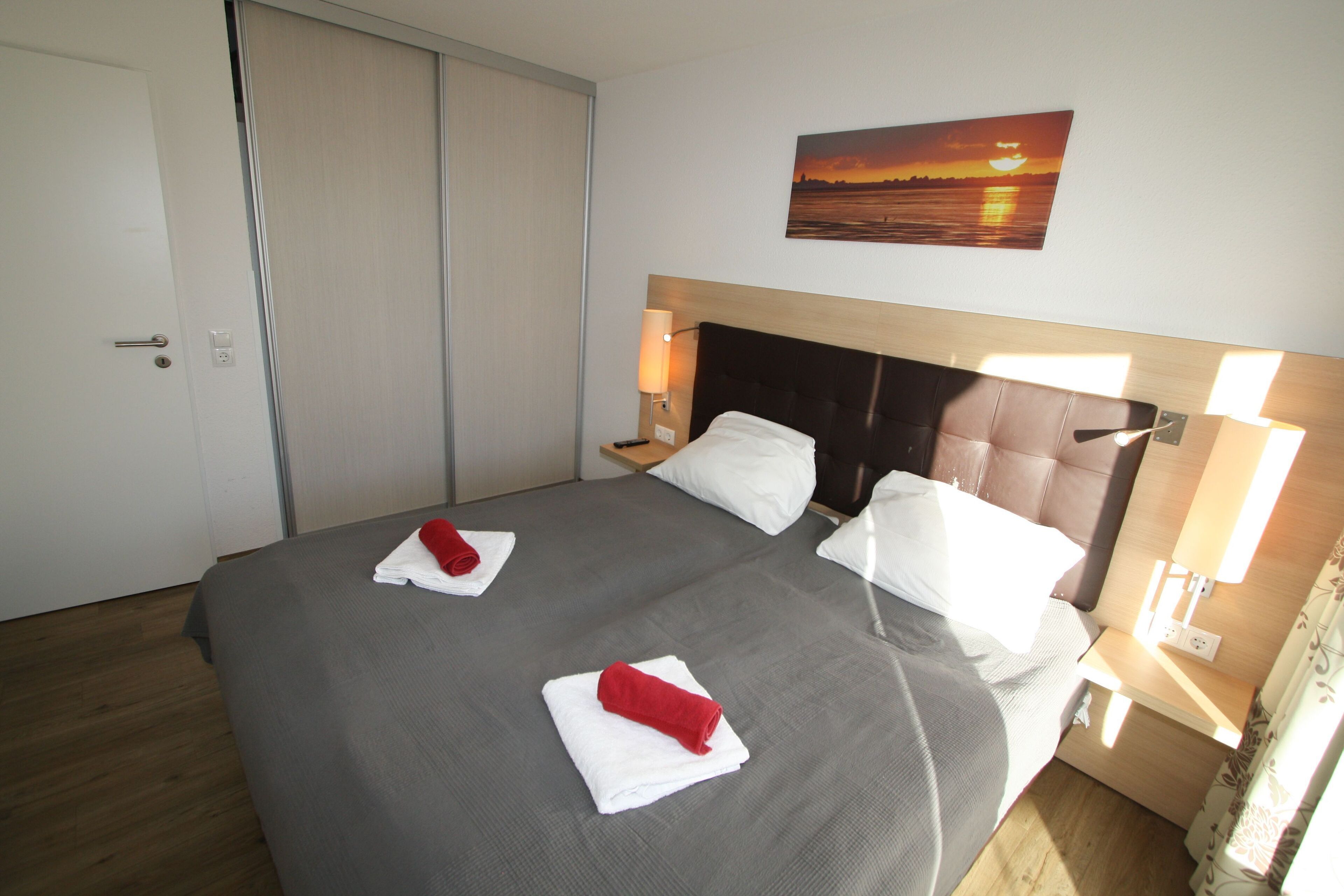 Une chambre d’hôtel avec un grand lit, deux oreillers, une table de chevet, une lampe fixée au mur et un tableau représentant un coucher de soleil.