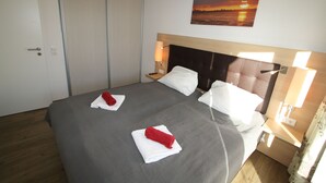 Una habitación de hotel con una cama grande, dos almohadas, una mesita de noche, una lámpara de pared y un cuadro de un atardecer.