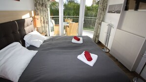 Habitación de hotel con una cama grande, dos almohadas blancas, una mesita de noche, una lámpara, un balcón con mesa y sillas, y vistas a zonas verdes.