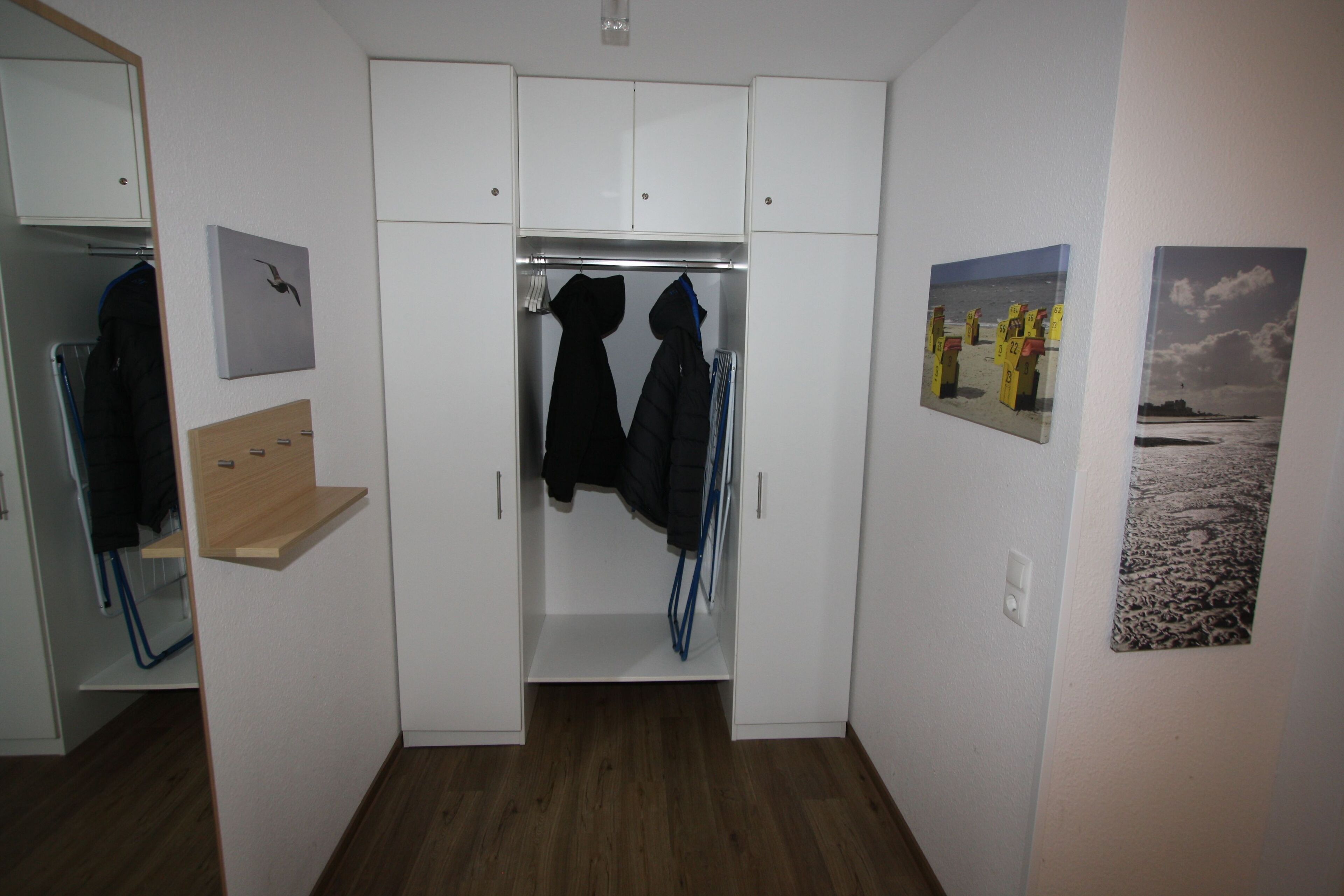 Un dressing accessible sans porte, avec un plancher en bois, des murs blancs et un miroir. Deux manteaux sont suspendus à un portant, et une étagère contient une veste.