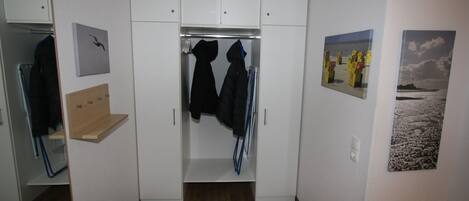 Un dressing accessible sans porte, avec un plancher en bois, des murs blancs et un miroir. Deux manteaux sont suspendus à un portant, et une étagère contient une veste.