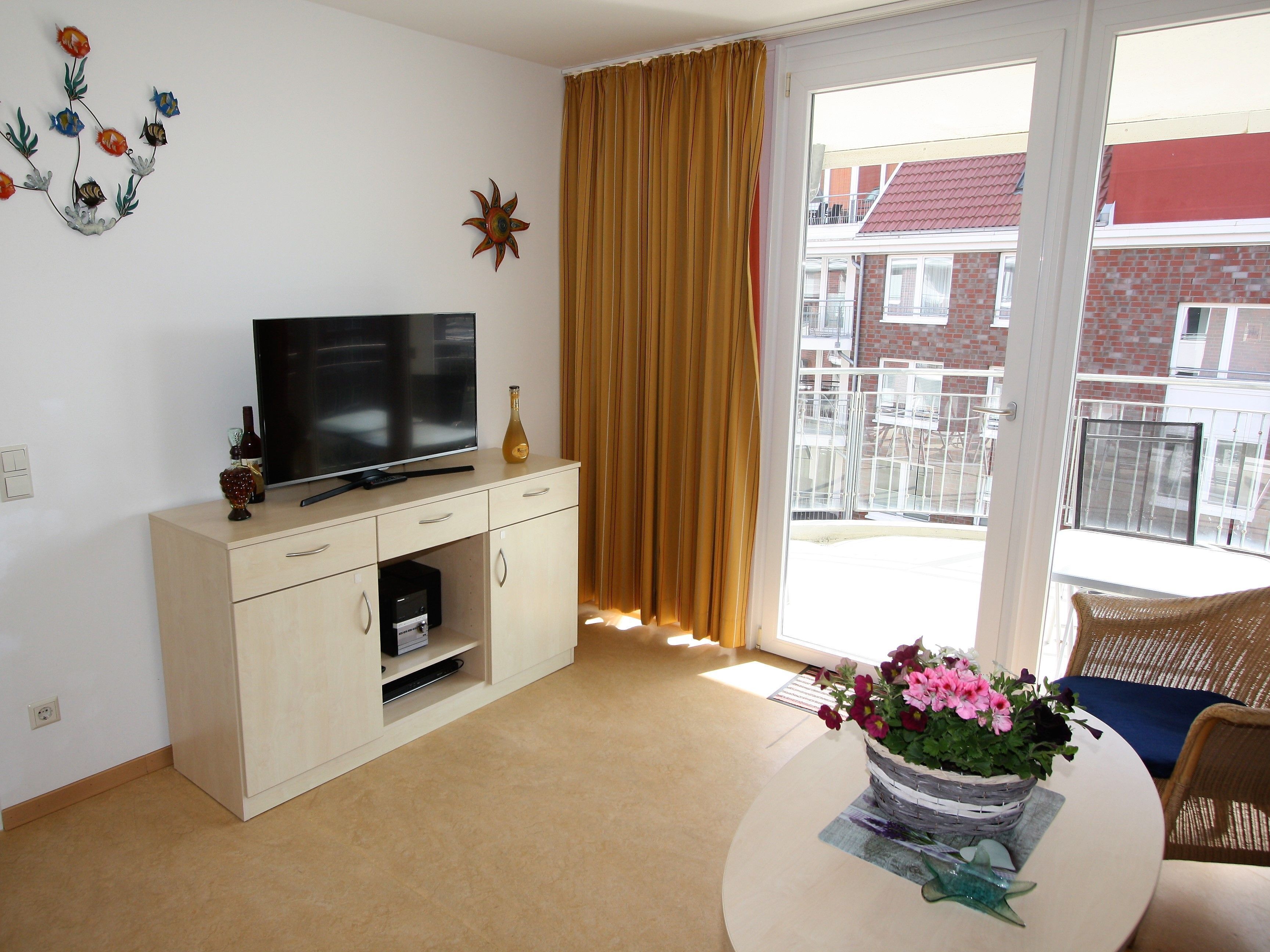 Una sala de estar con un televisor sobre un mueble bajo, una ventana con cortinas, un balcón con mobiliario exterior y una mesa con un jarrón de flores.