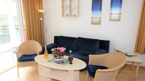 Een woonkamer met een donkerblauwe bank, twee rieten fauteuils, een witte salontafel met een fles en bloemen, een klein witte tafeltje, en een wandplank met decoratieve objecten.