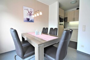 Un coin repas avec une table en bois, des chaises noires, un set de table rose et un vase avec des fleurs accroché au mur.
