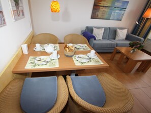 A dining area with a table set for two, two wicker chairs, a grey sofa, and a small side table with a vase.
