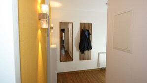 A hallway with a wooden floor, a coat rack with jackets, a mirror, and a light fixture.