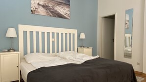 Ein Schlafzimmer mit einem weißen Bett, zwei Nachttischen mit Lampen, einem großen Gemälde an der Wand und einer Tür zu einem anderen Raum.